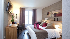 A hotel room with a bed, bedside table, desk, chair, TV, window with curtains, and artwork on the wall.