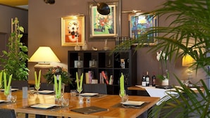 A dining area with a long wooden table set for a meal, flanked by chairs. There are framed artworks on the wall, a bookshelf with books and decorative items, and a potted plant in the foreground.