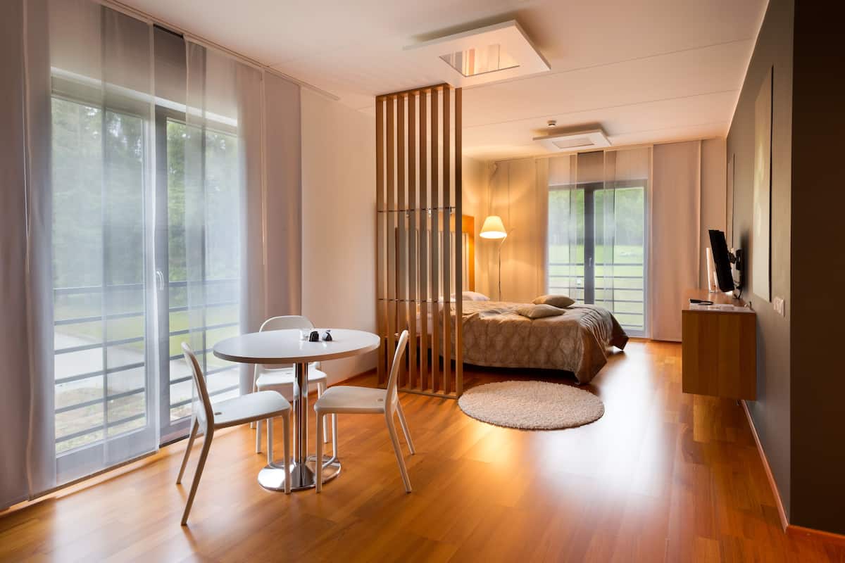 A modern hotel room with a large window, a round dining table with chairs, a bed with a headboard, and a small bedside table.