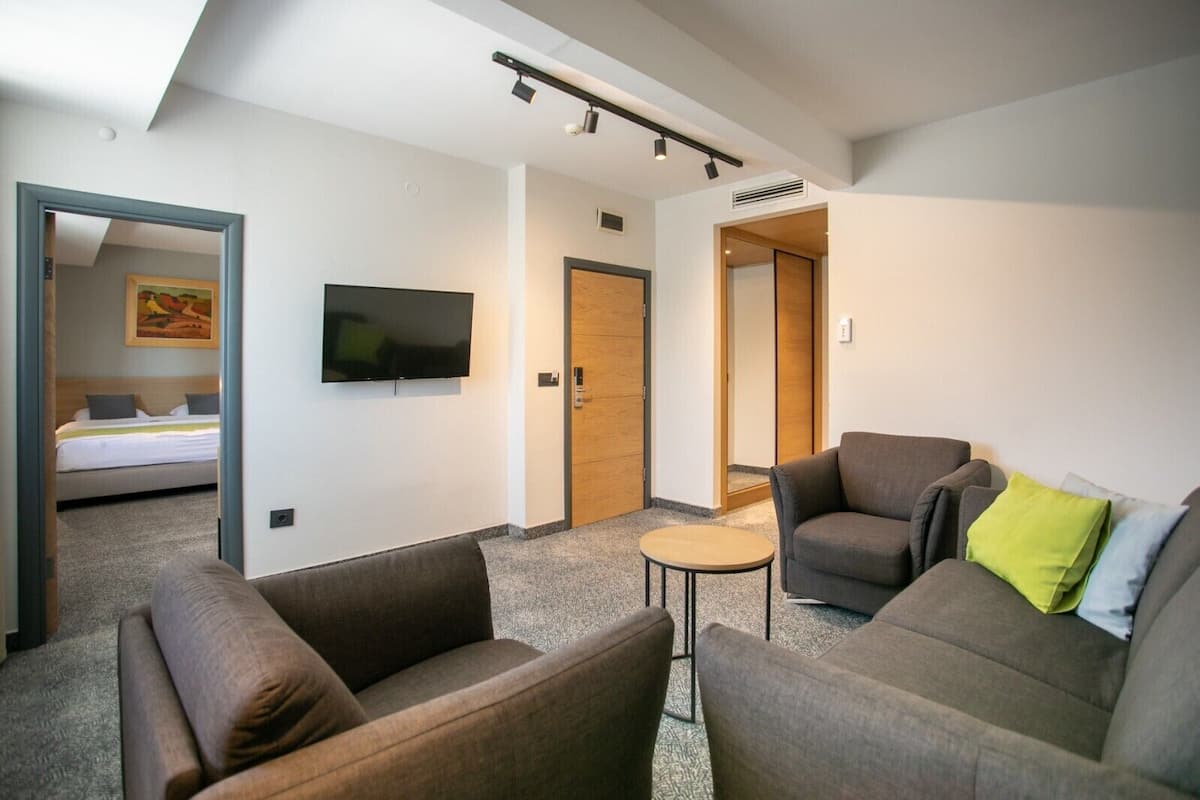A modern hotel room with a large bed, a flat-screen TV, two sofas, a small round table, and a wooden door.