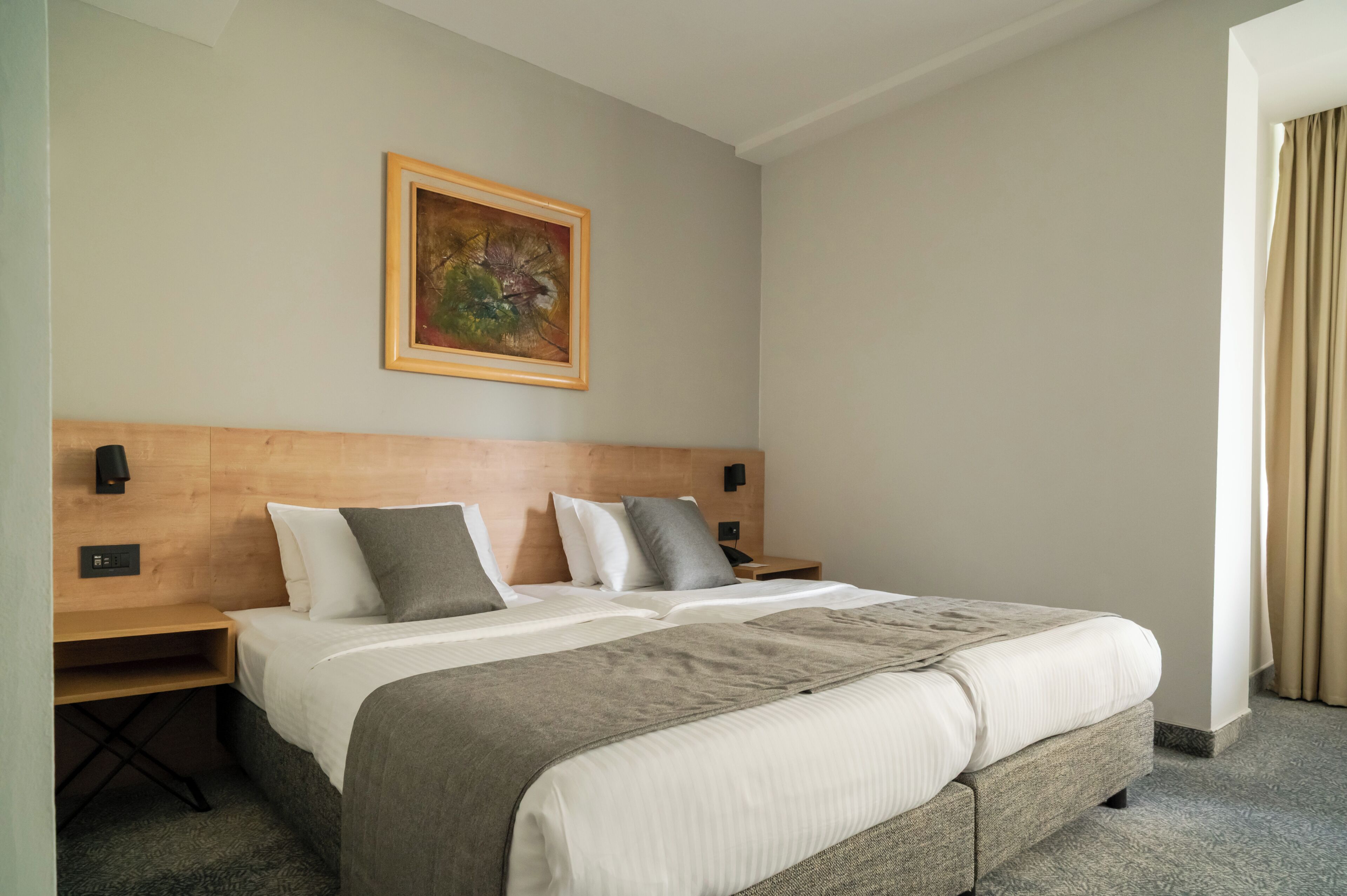 A hotel room with a bed, a nightstand, a painting on the wall, and a window with curtains.