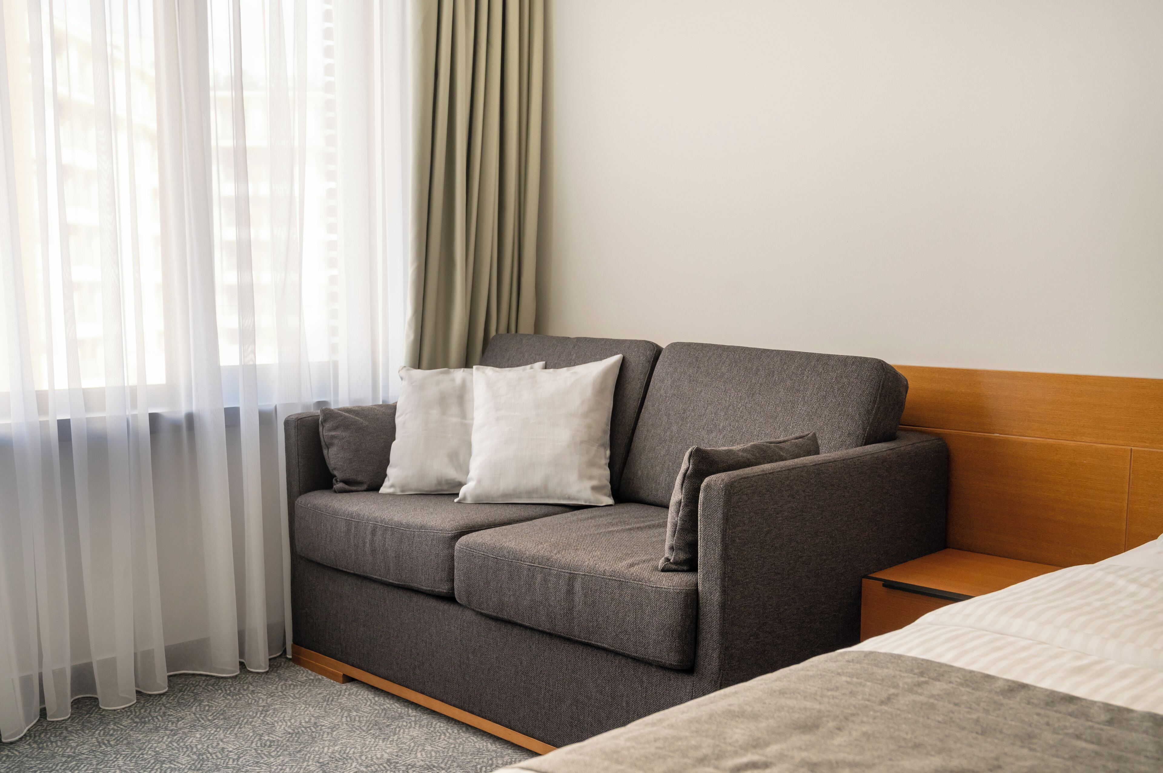 A modern hotel room with a grey sofa, a wooden headboard, and sheer curtains.