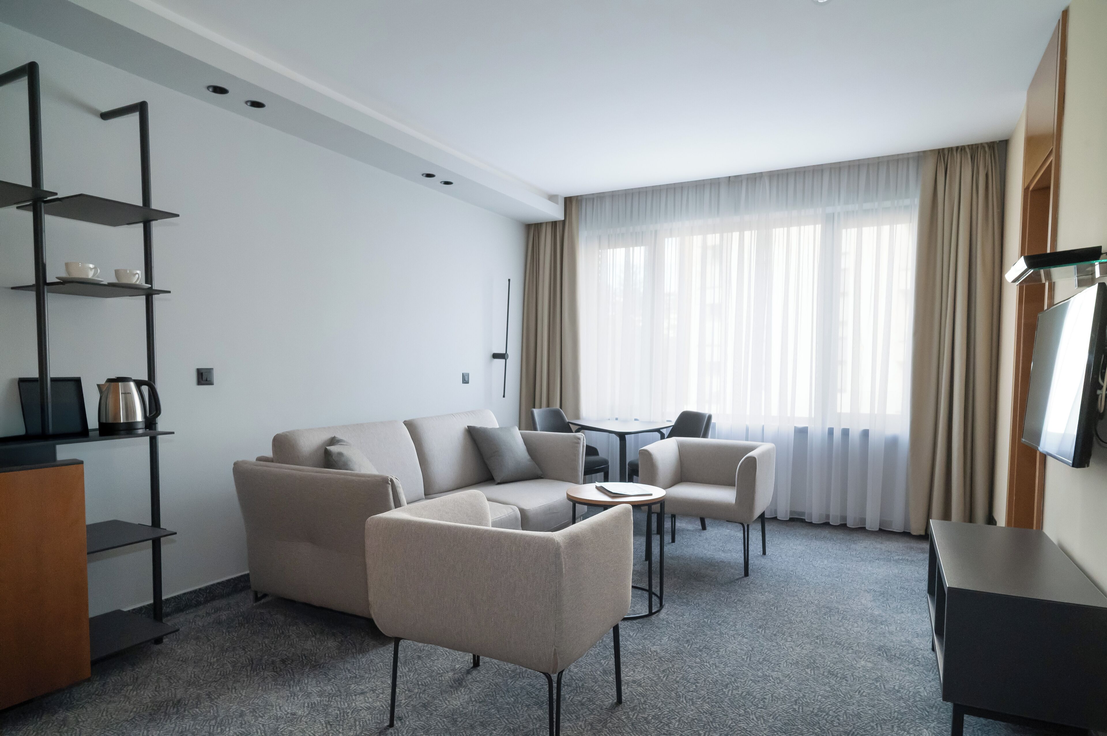 Una habitación de hotel moderna con un sofá seccional, una pequeña mesa redonda, un televisor y una estantería.