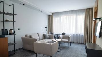 Ein modernes Hotelzimmer mit einer Eckcouch, einem kleinen runden Tisch, einem Fernseher und einem Bücherregal.