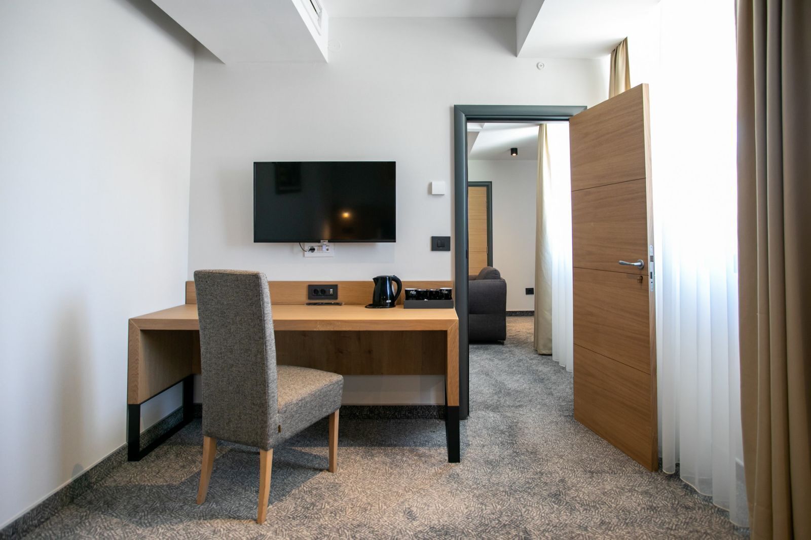 Une chambre d’hôtel moderne avec un bureau en bois, un téléviseur à écran plat, un fauteuil rembourré de couleur grise et un sol recouvert de moquette.