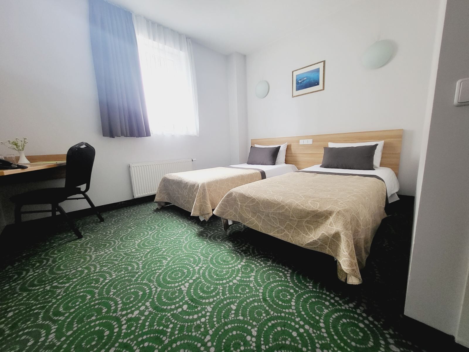 Une chambre d’hôtel avec deux lits, un bureau, une chaise, un radiateur, une fenêtre avec des rideaux, une horloge murale et un tableau encadré.
