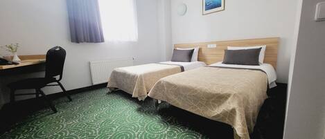 Une chambre d’hôtel avec deux lits, un bureau, une chaise, un radiateur, une fenêtre avec des rideaux, une horloge murale et un tableau encadré.