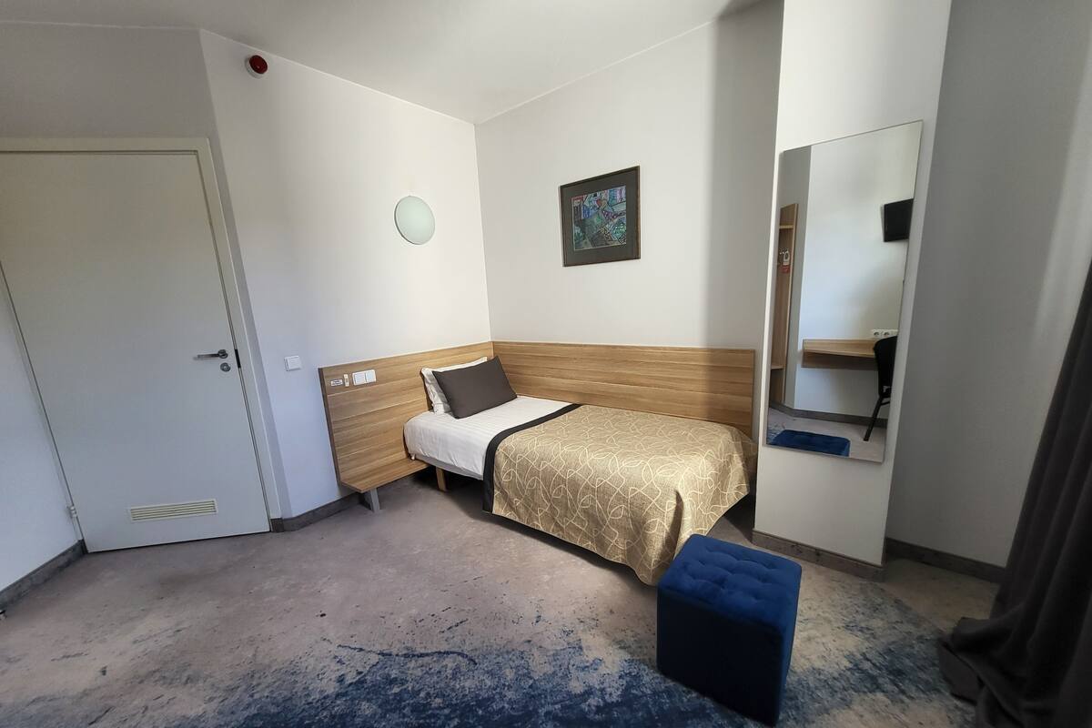 Una habitación de hotel con cama, un taburete pequeño, un espejo y una puerta.
