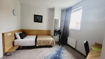 A small, single-room hotel room with a bed, a desk, a chair, a radiator, a window with curtains, and a wall-mounted fan.