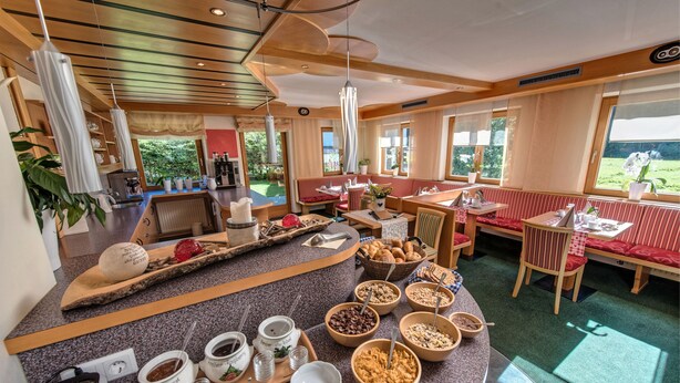 Un bufé de desayuno con diversos alimentos y bebidas, una zona de estar con bancos rojos acolchados y vistas al exterior con zonas verdes.