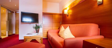 A modern hotel room with a red sofa, a flat-screen TV mounted on the wall, a wooden accent wall, and a red carpet.