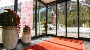 Un hall de hotel con ventanas amplias, una alfombra roja y dos grandes macetas con plantas.