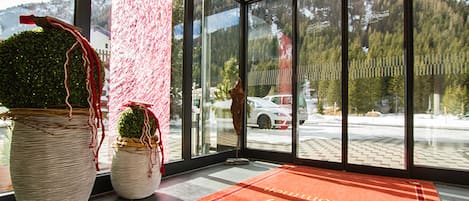 Un hall de hotel con ventanas amplias, una alfombra roja y dos grandes macetas con plantas.
