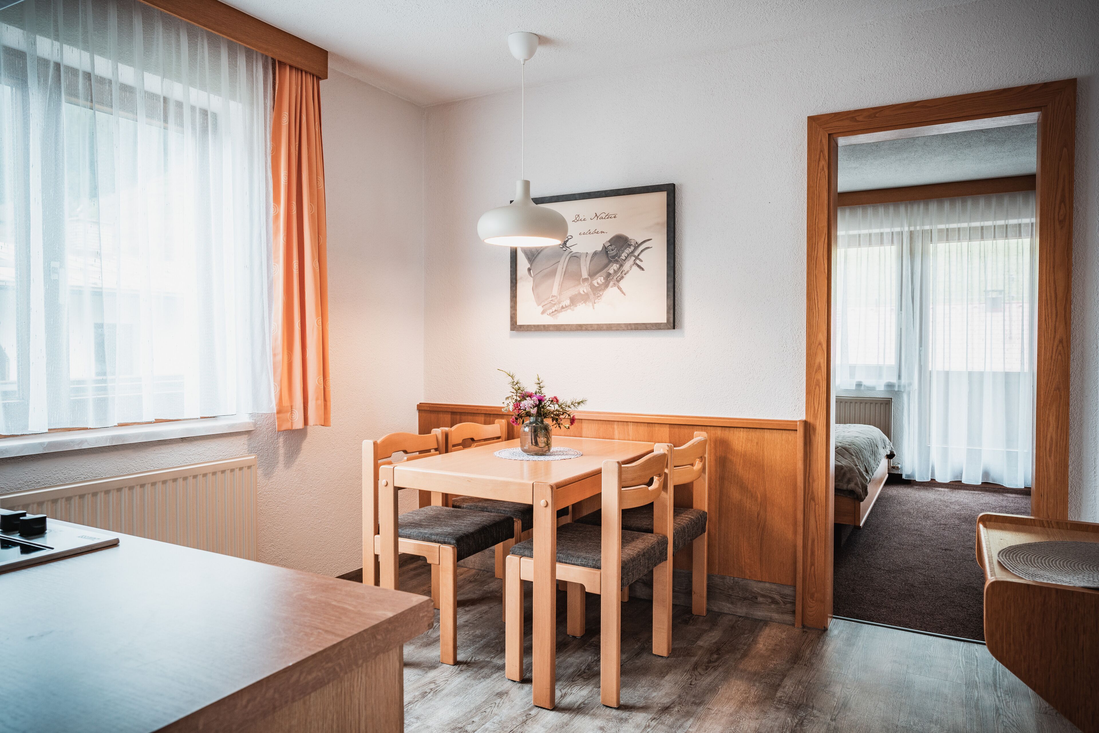 A hotel room with a wooden dining table, four chairs, a small side table, a framed picture on the wall, and a view of the adjacent bedroom through an open door.
