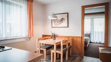 A hotel room with a wooden dining table, four chairs, a small side table, a framed picture on the wall, and a view of the adjacent bedroom through an open door.