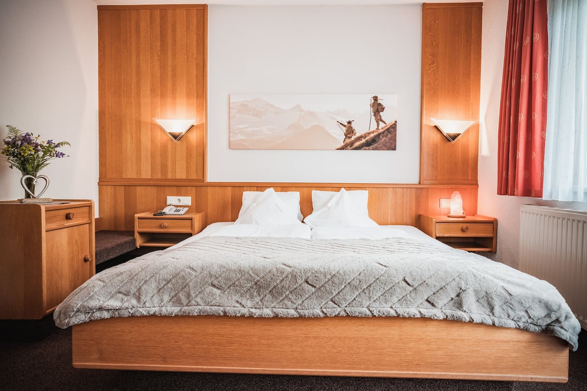 Une chambre d’hôtel avec un lit en bois, deux tables de chevet, une lampe fixée au mur, un tableau encadré et un vase avec des fleurs.