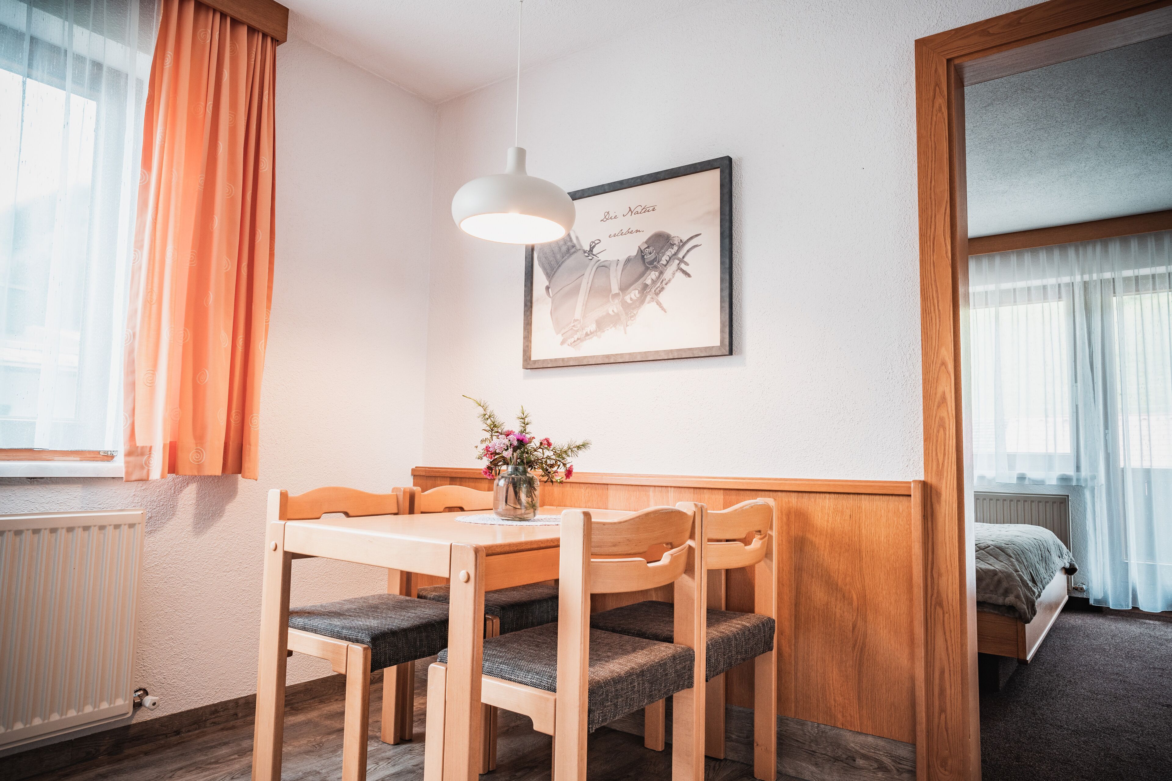 A room with a wooden dining table, four chairs, a framed picture on the wall, a window with orange curtains, and a radiator.