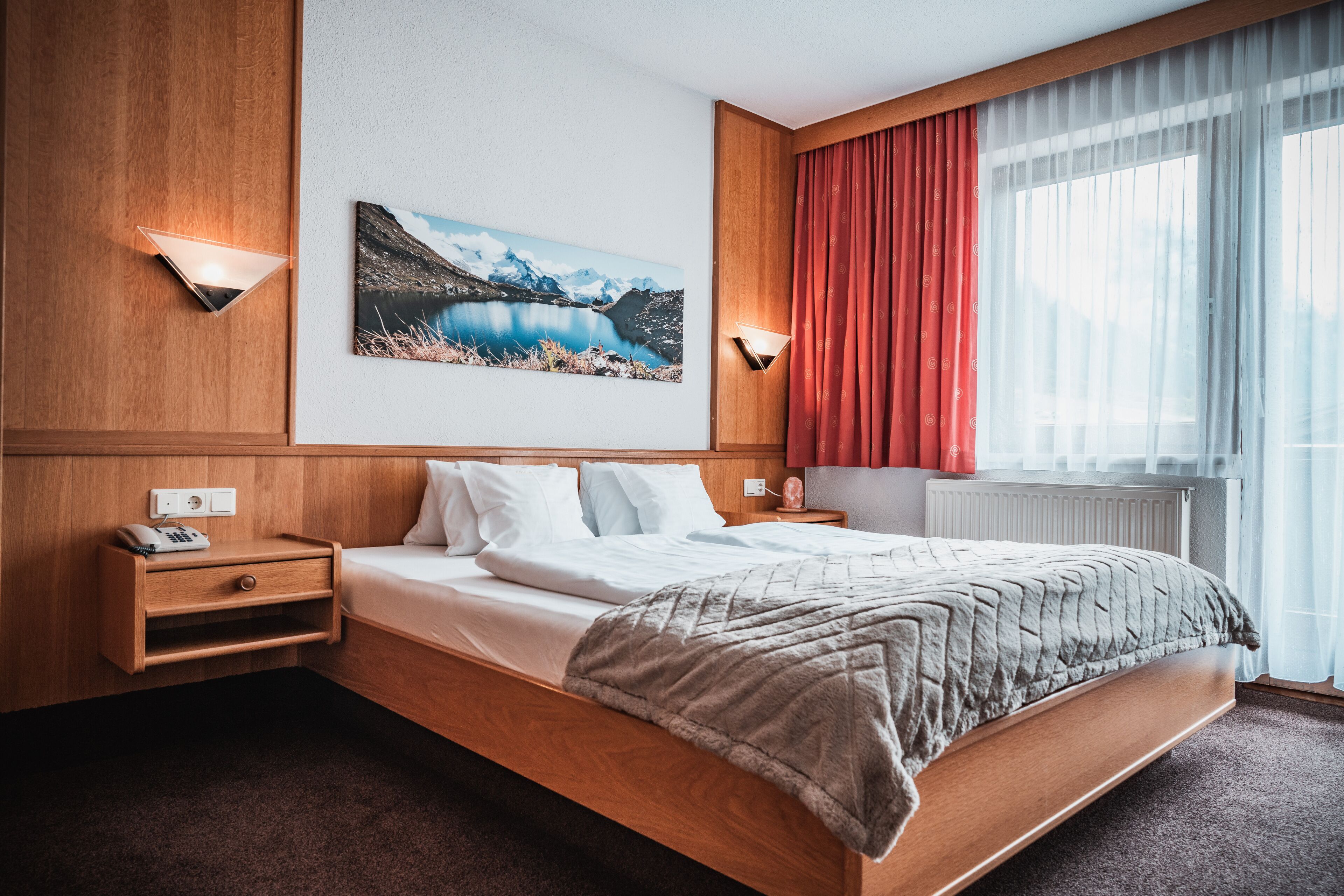 Una habitación de hotel con una cama de madera, una mesita de noche con un teléfono, un cuadro de un lago y una ventana con cortinas rojas.