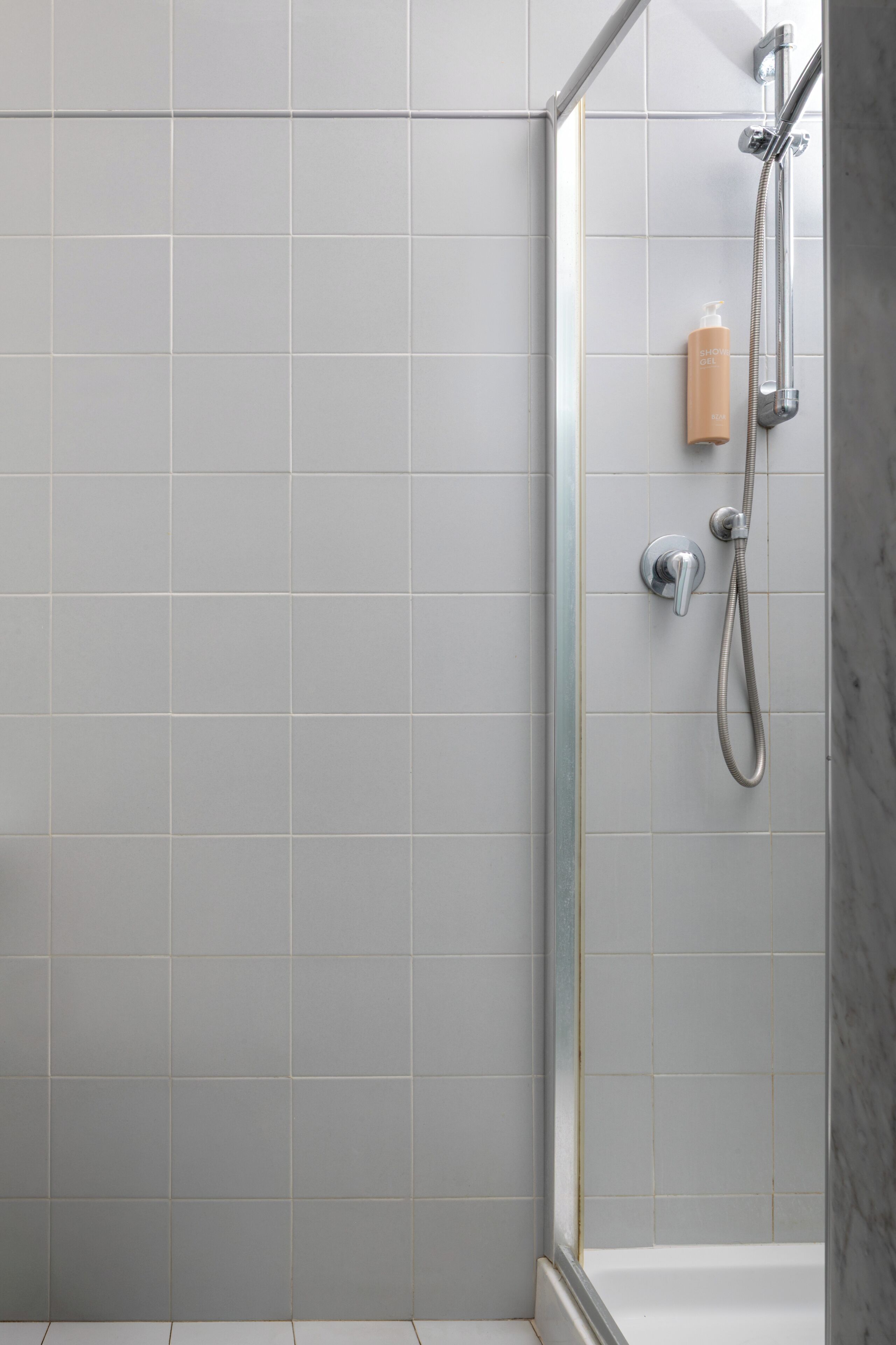 Area doccia con porta in vetro, soffione a mano e flacone di shampoo.