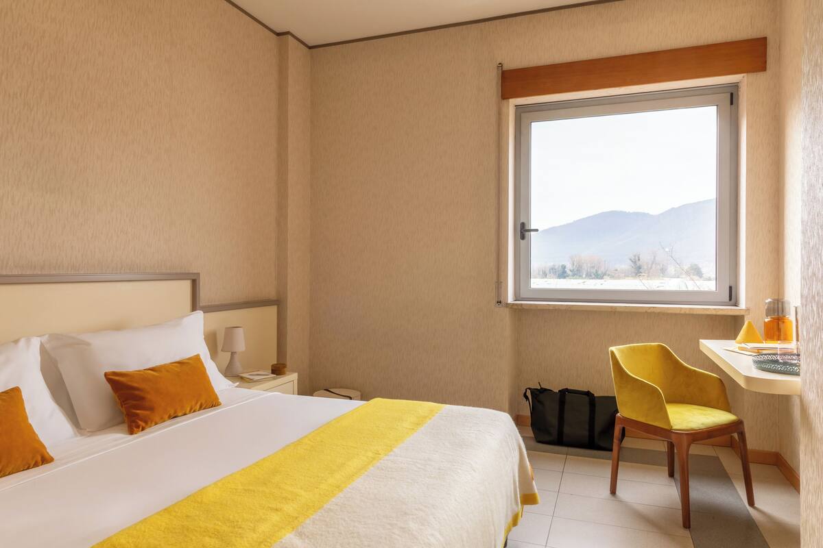 Une chambre d’hôtel avec un lit, un bureau, une chaise, une fenêtre donnant sur la montagne et une valise posée sur le sol.