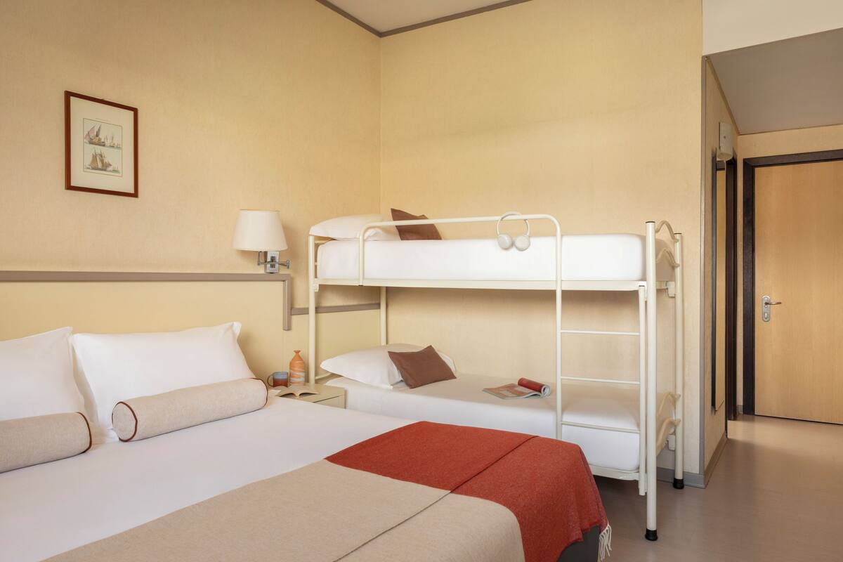 Une chambre d’hôtel avec des lits superposés, une table de chevet avec une lampe, un tableau encadré au mur et une porte sur la droite.