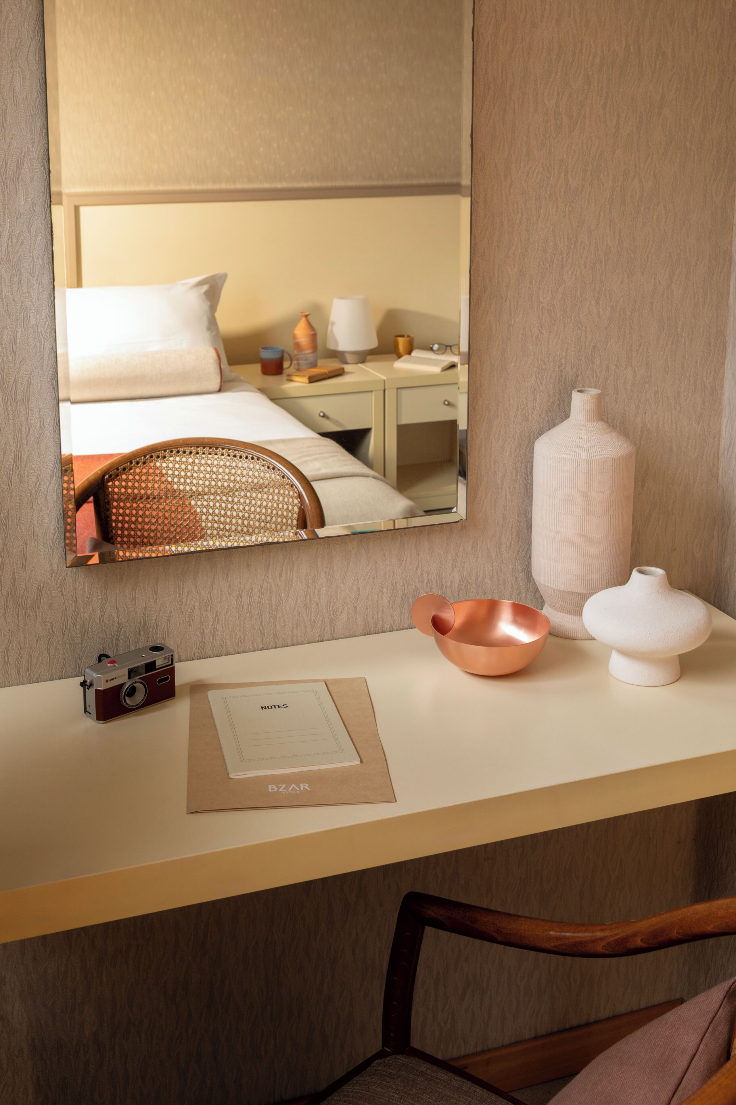 A hotel room with a bed, a desk with a camera, a vase, and a mirror reflecting the room.