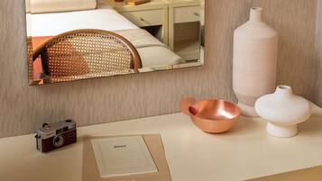 A hotel room with a bed, a desk with a camera, a vase, and a mirror reflecting the room.