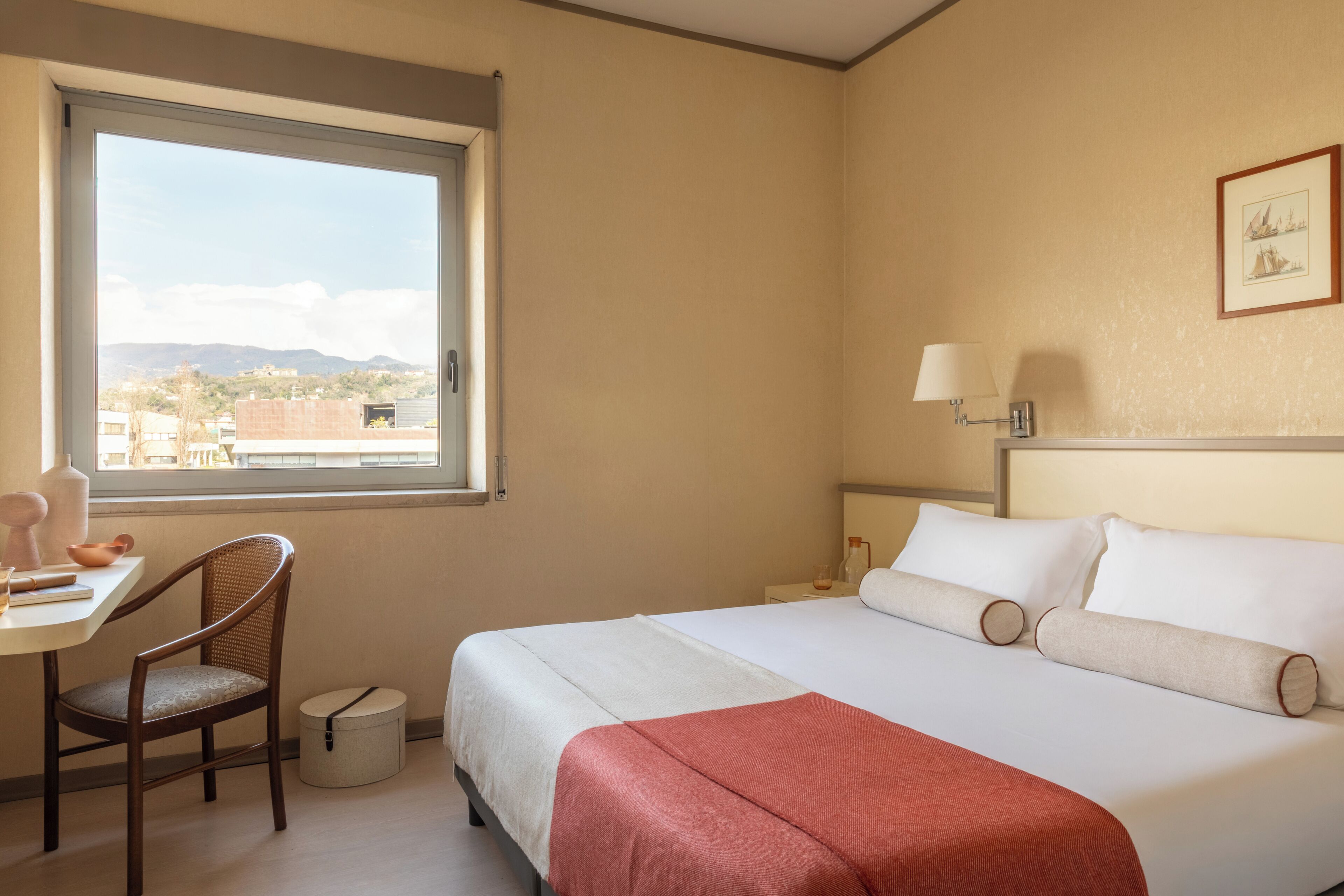 Una habitación de hotel con cama, escritorio, silla, ventana y lámpara fijada a la pared.