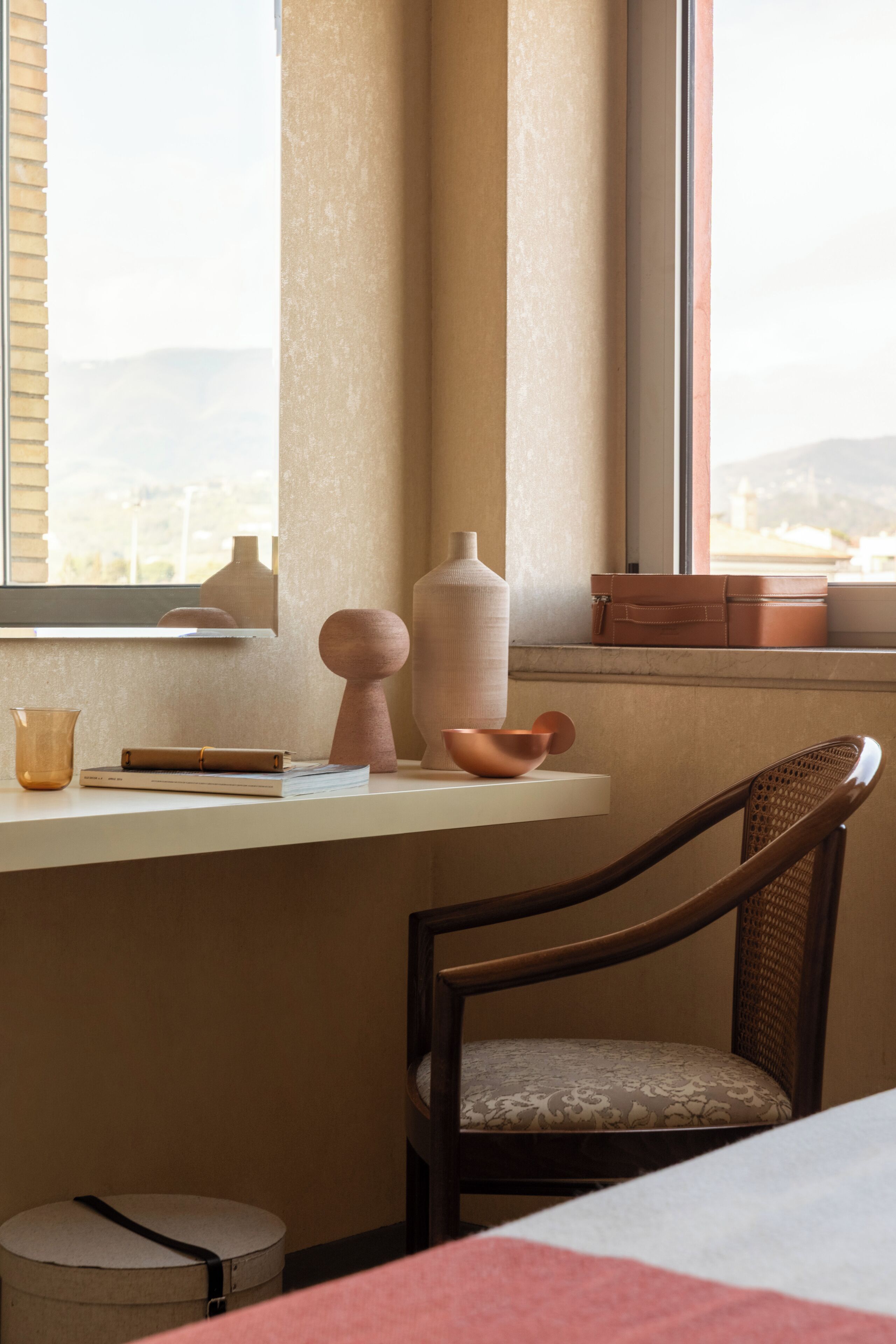 Une pièce avec une fenêtre, un bureau sur lequel est posé un vase, une chaise et un petit pouf rond.