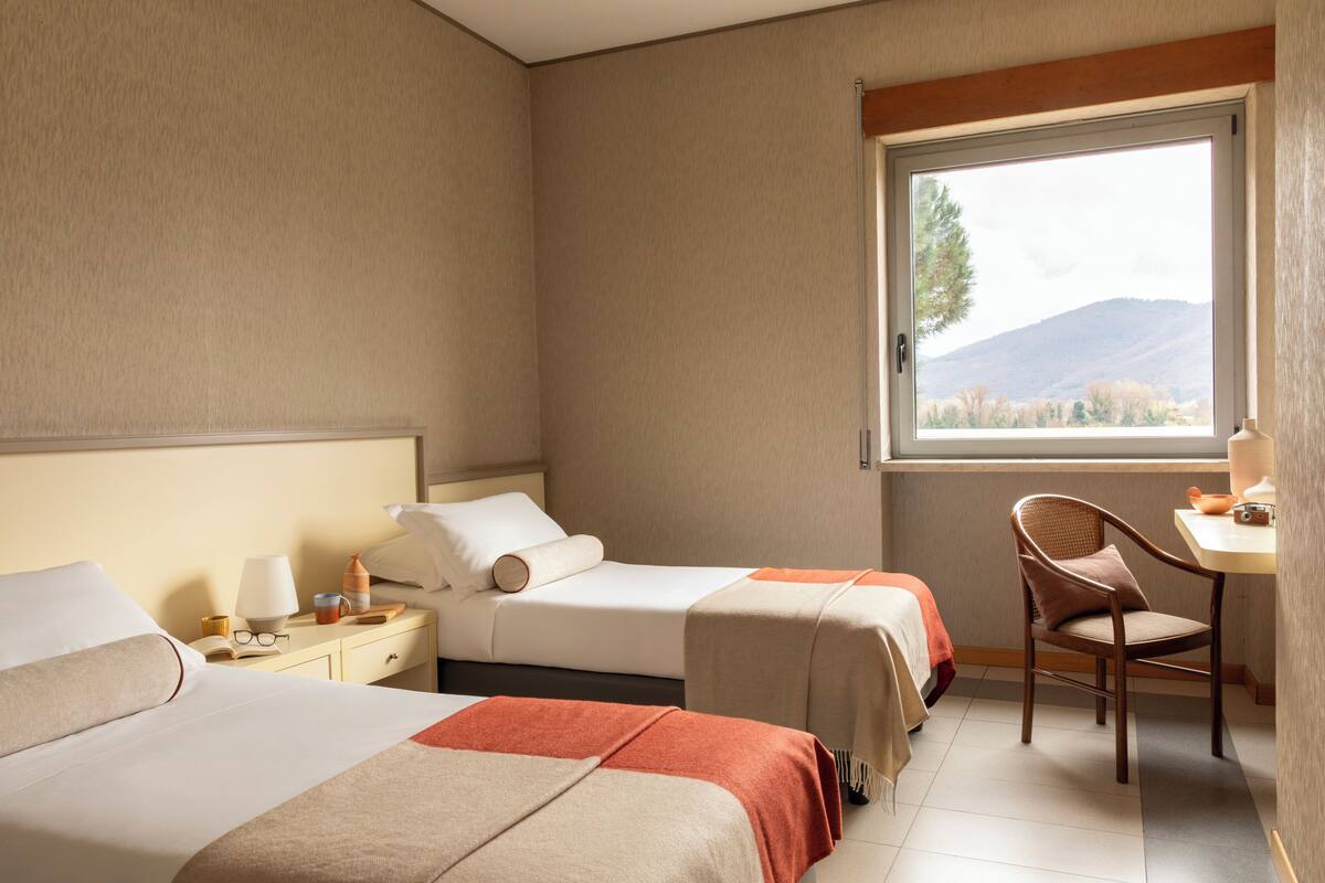 Une chambre d’hôtel avec deux lits, un bureau, une chaise et une fenêtre donnant sur des collines.