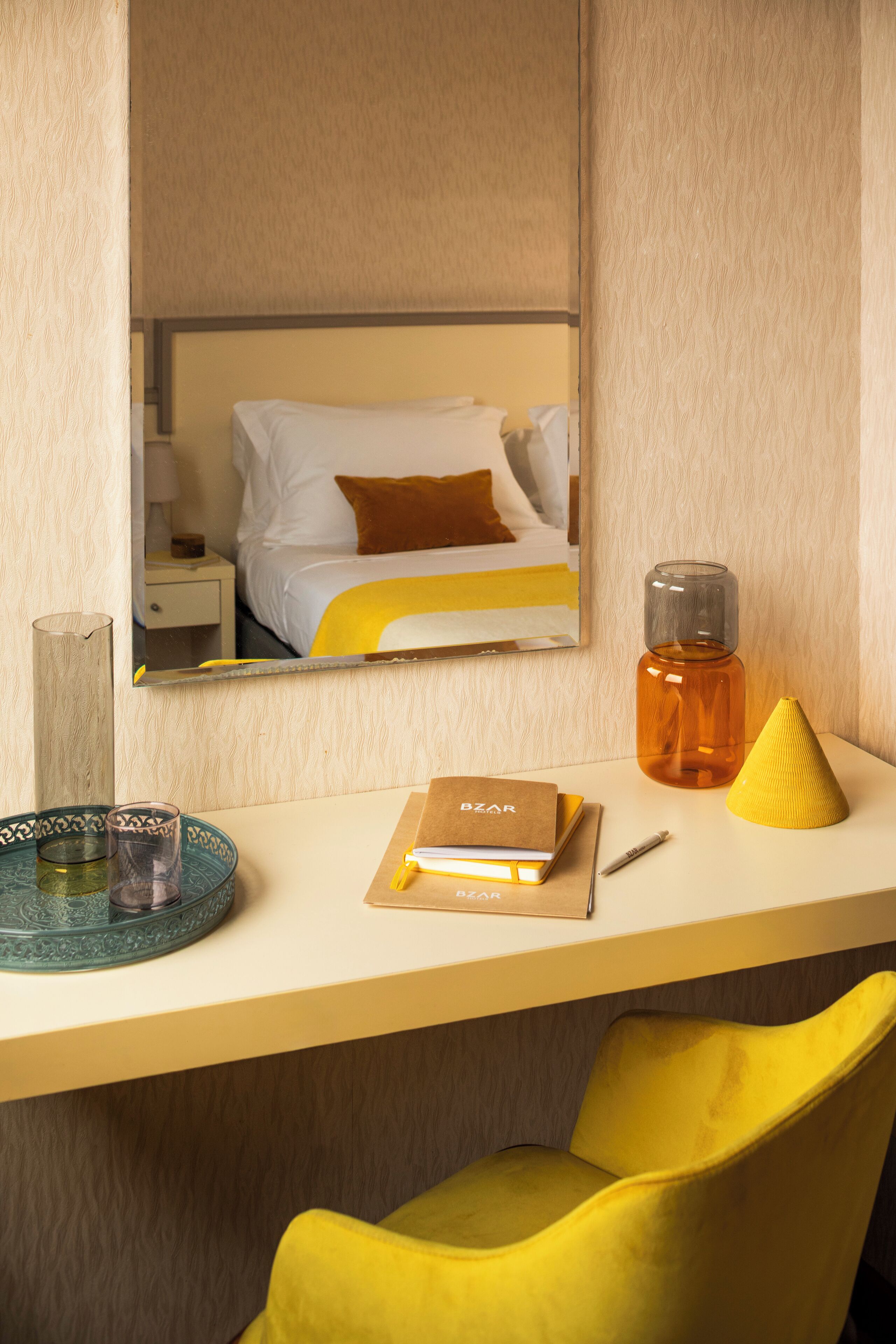 Ein Hotelzimmer mit einem Bett, einem Nachttisch mit Lampe, einem Spiegel, einem kleinen Tisch mit einem Buch, einer Flasche und einem gelben Stuhl.
