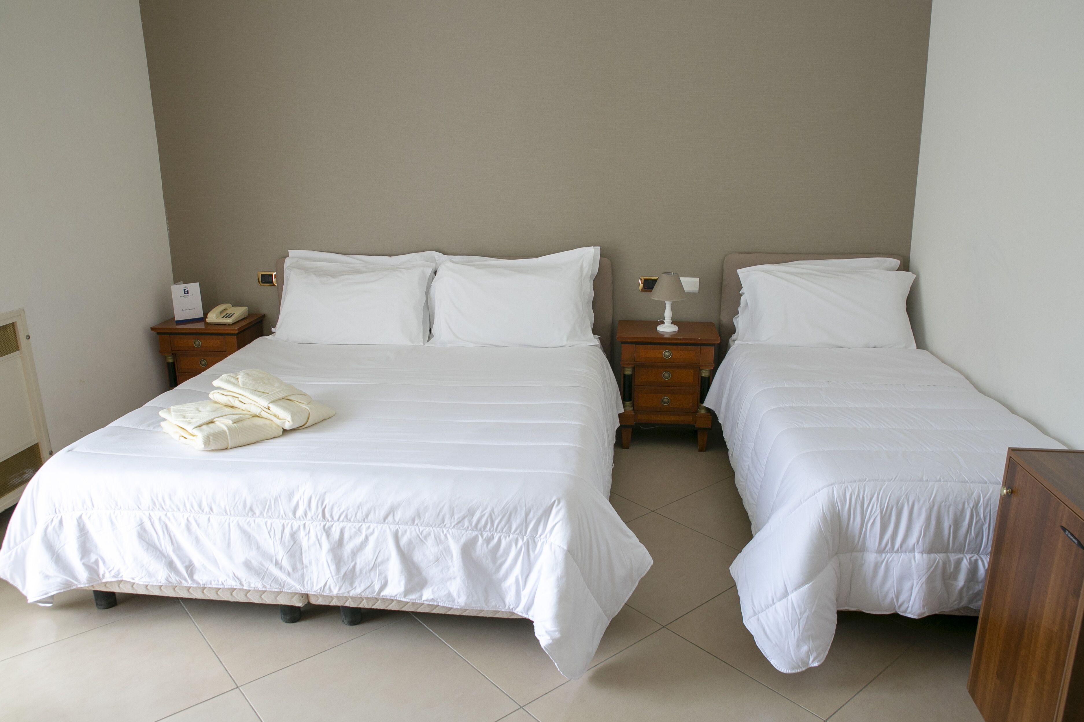 A double bed with white bedding and a folded towel on the left side. There are two bedside tables, each with a lamp and a phone.