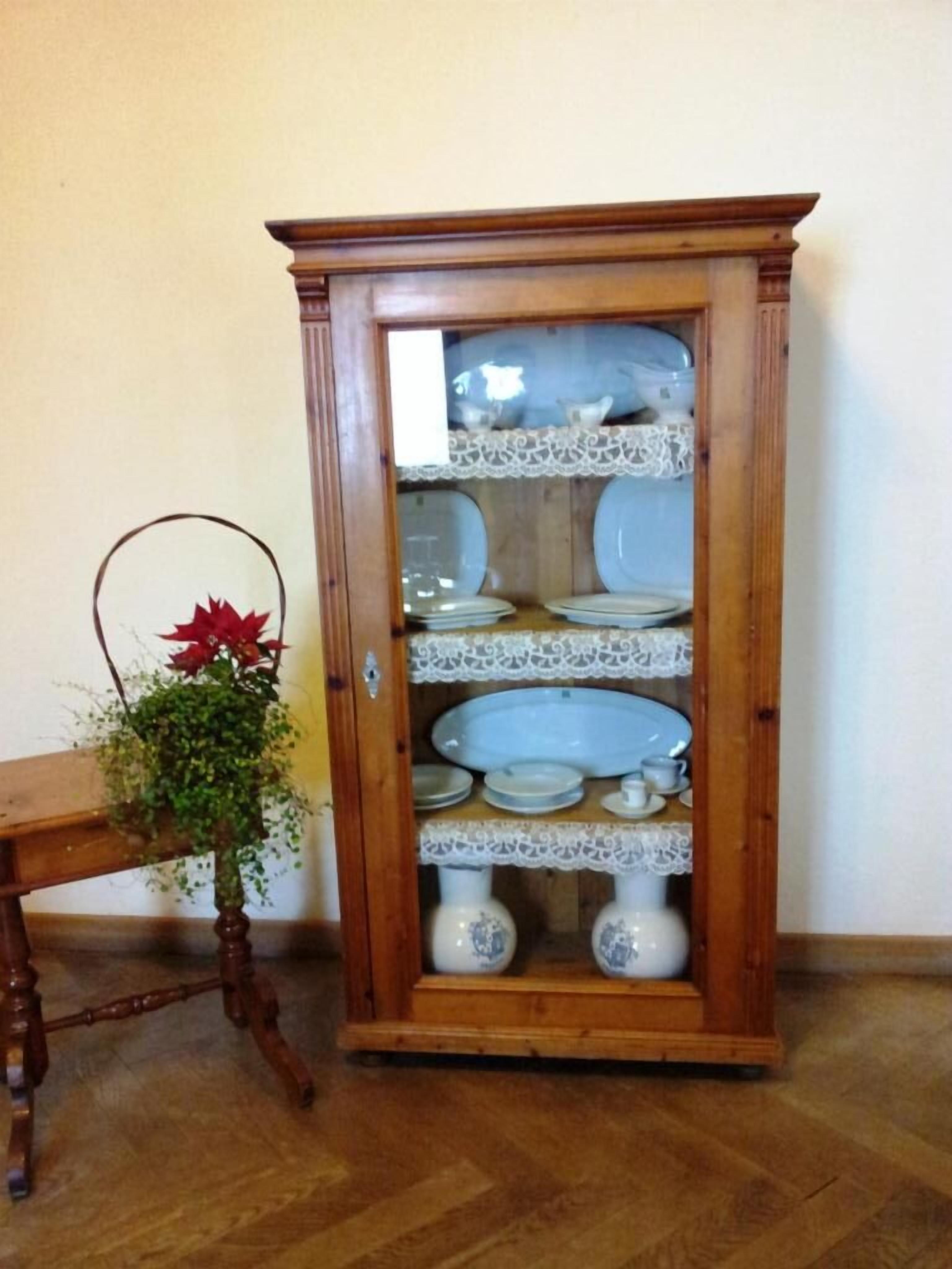 A wooden china cabinet with multiple shelves, a lace-trimmed top, and a glass door.