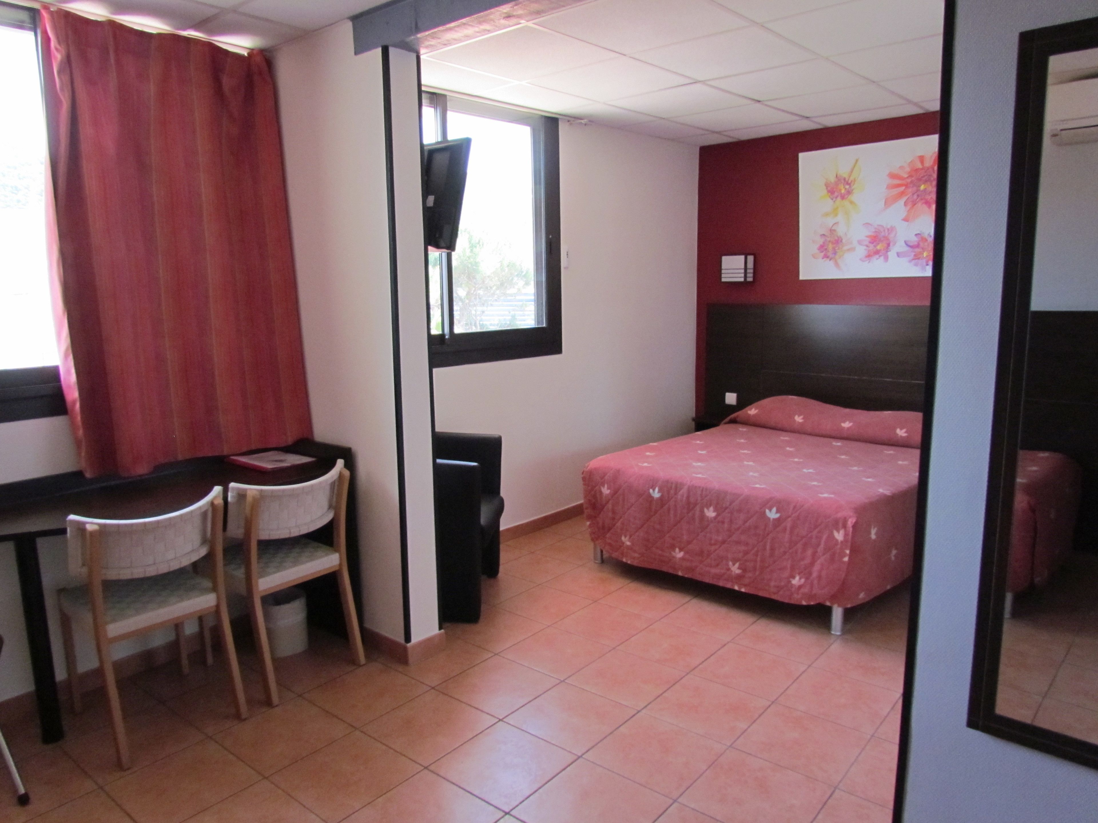 A bedroom with a bed, a desk, two chairs, a window with curtains, a TV, and a wall with a floral painting.