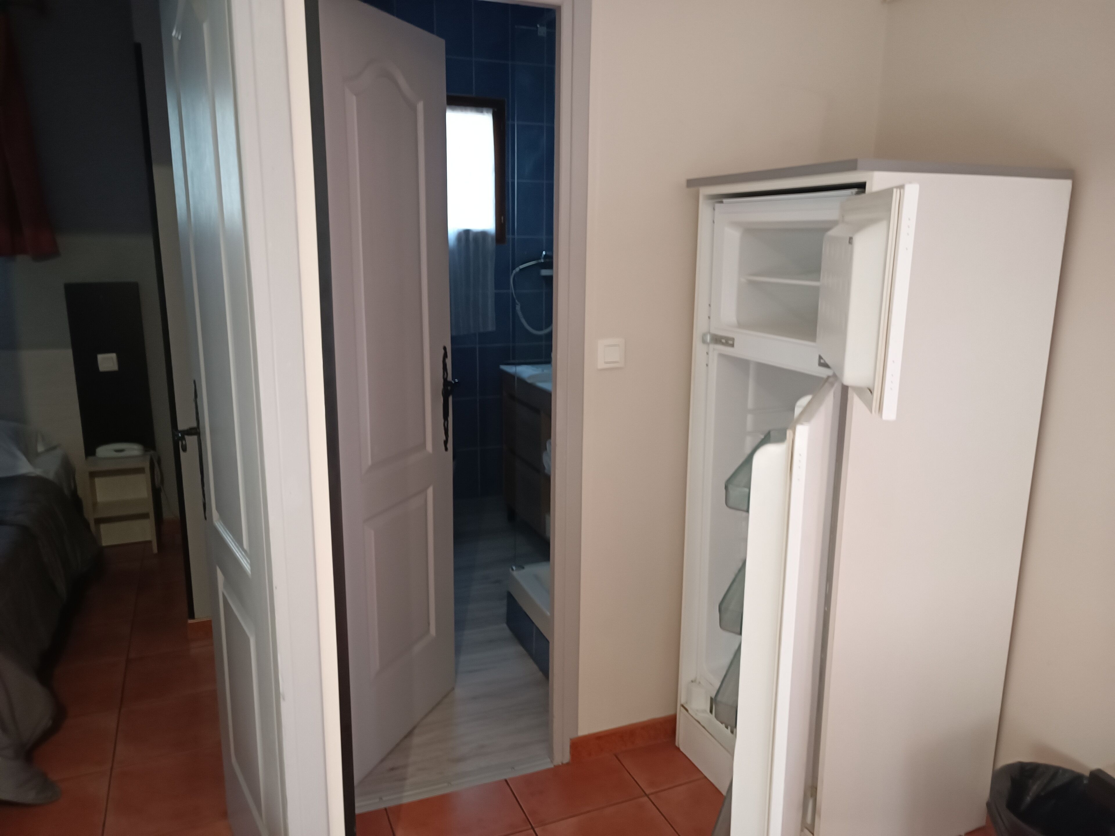 Una habitación con una puerta blanca que da a un baño, un refrigerador blanco y una cama con una manta gris.