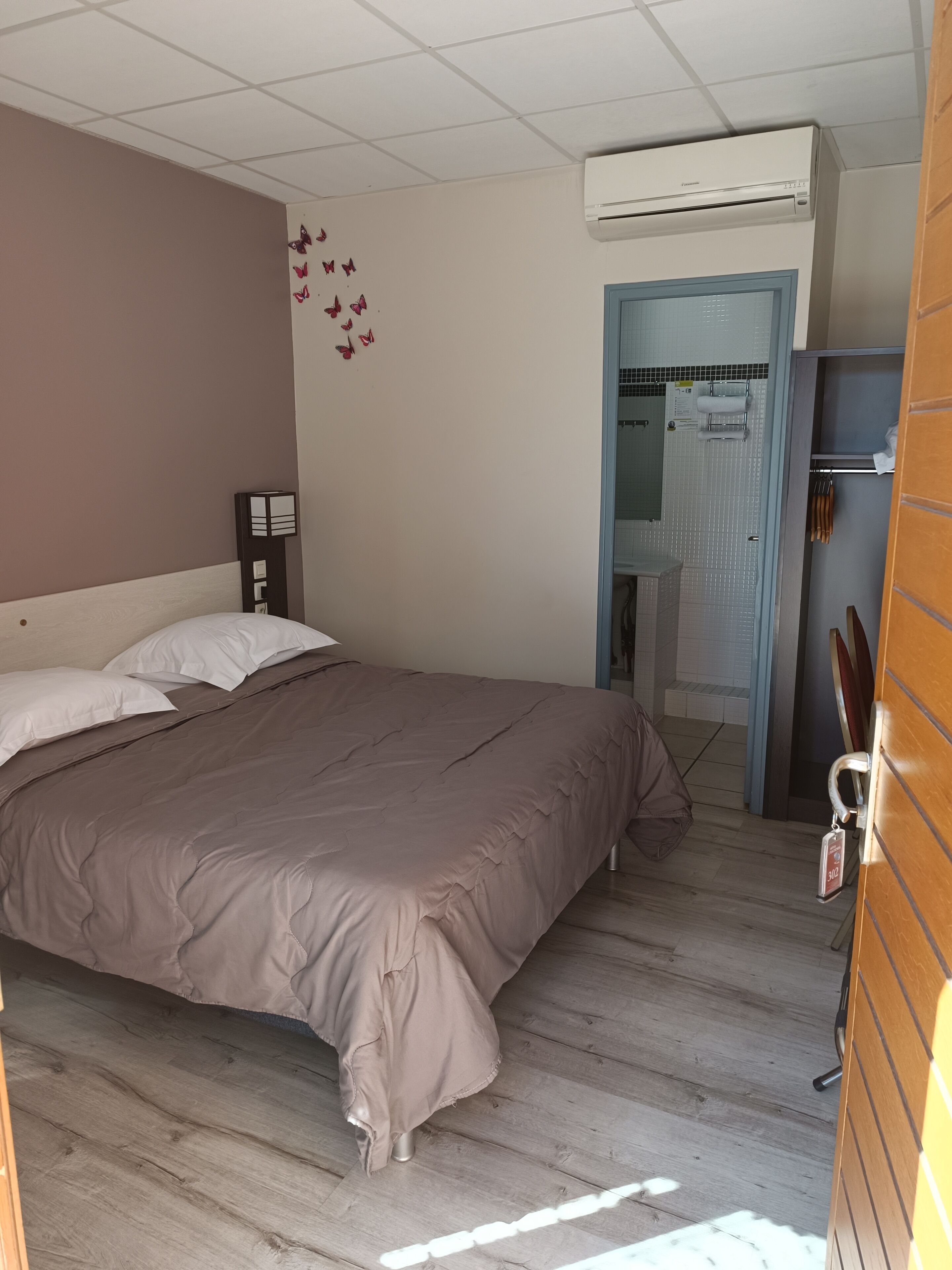 A bedroom with a large bed, an air conditioning unit, a wooden floor, and a bathroom visible through an open door.