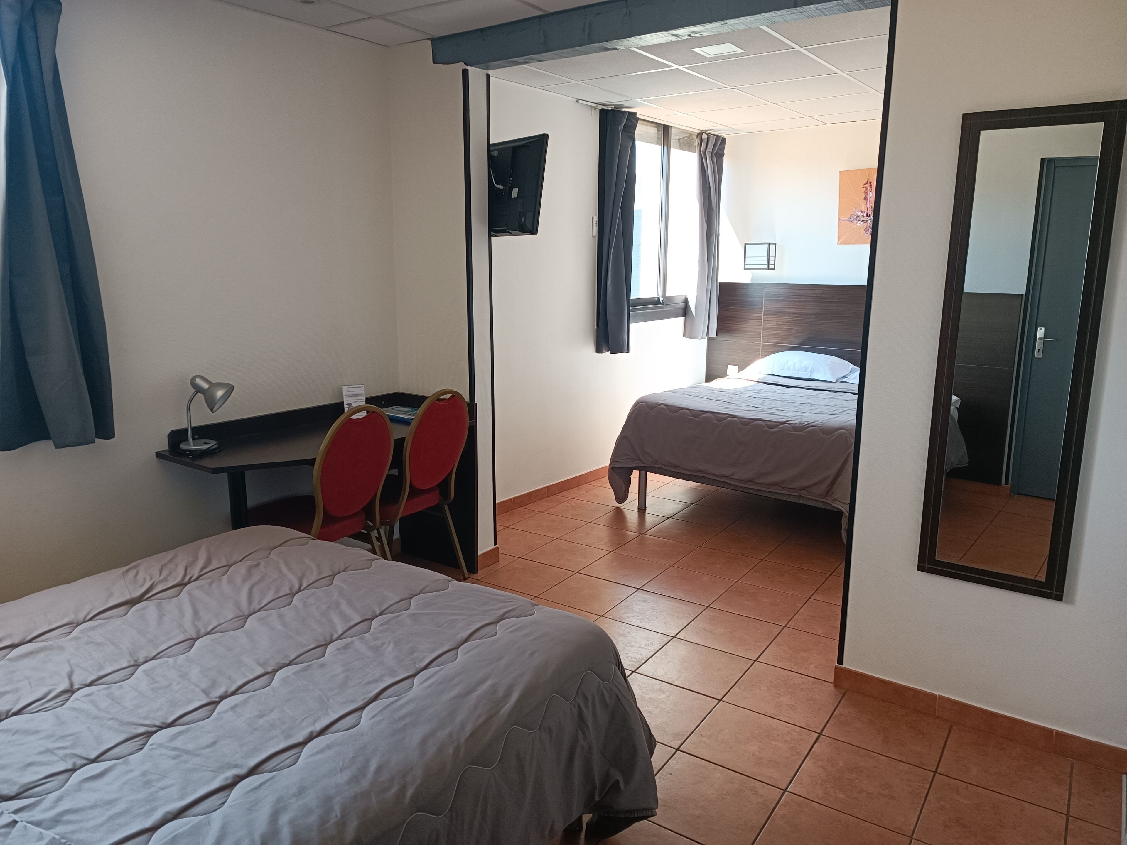 A hotel room with a bed, a desk with a chair, a window with curtains, and a mirror on the wall.