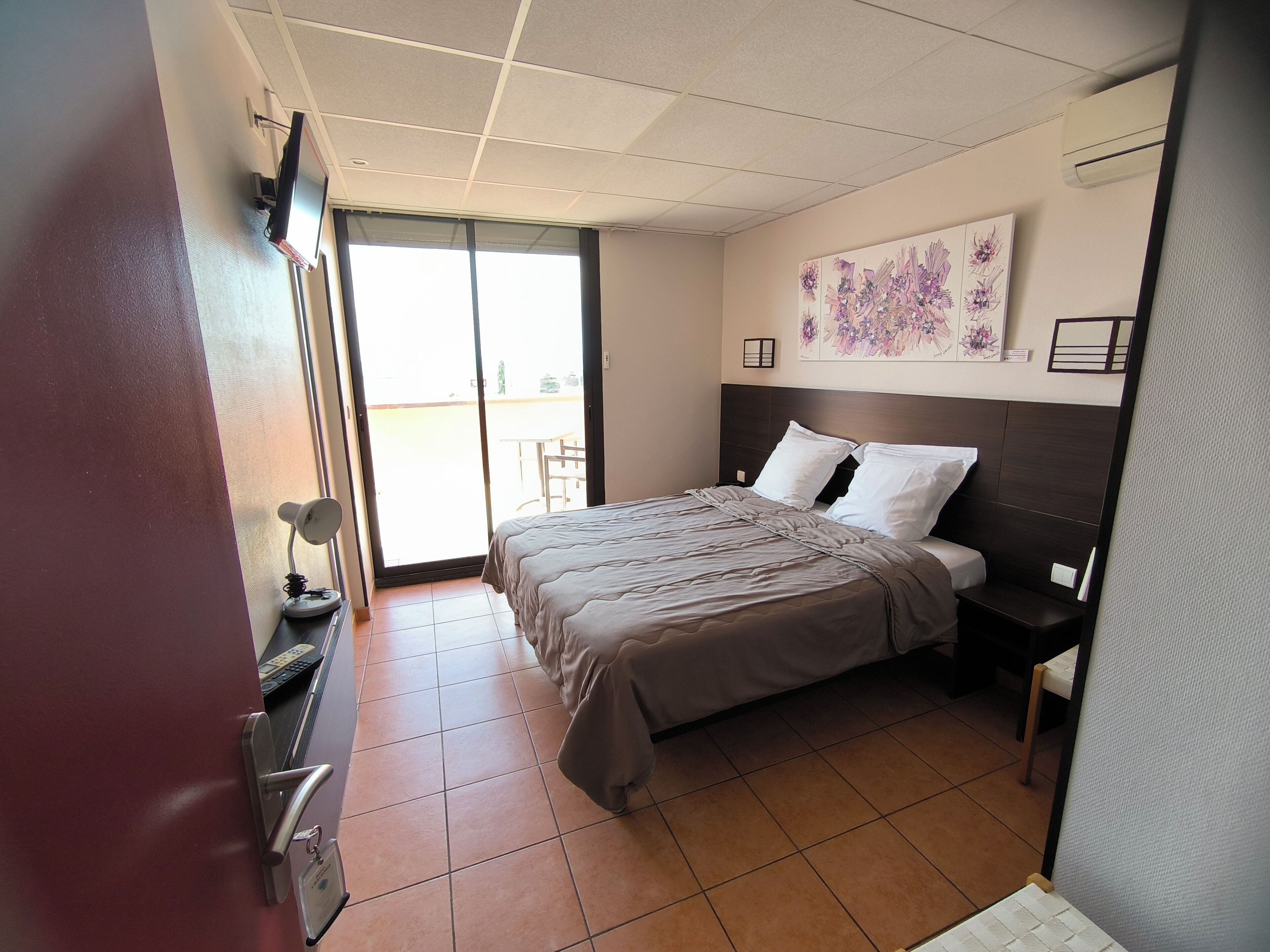 Una camera d'albergo con un letto, una scrivania, una sedia, una TV e una porta scorrevole su un balcone.