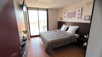 Una camera d'albergo con un letto, una scrivania, una sedia, una TV e una porta scorrevole su un balcone.