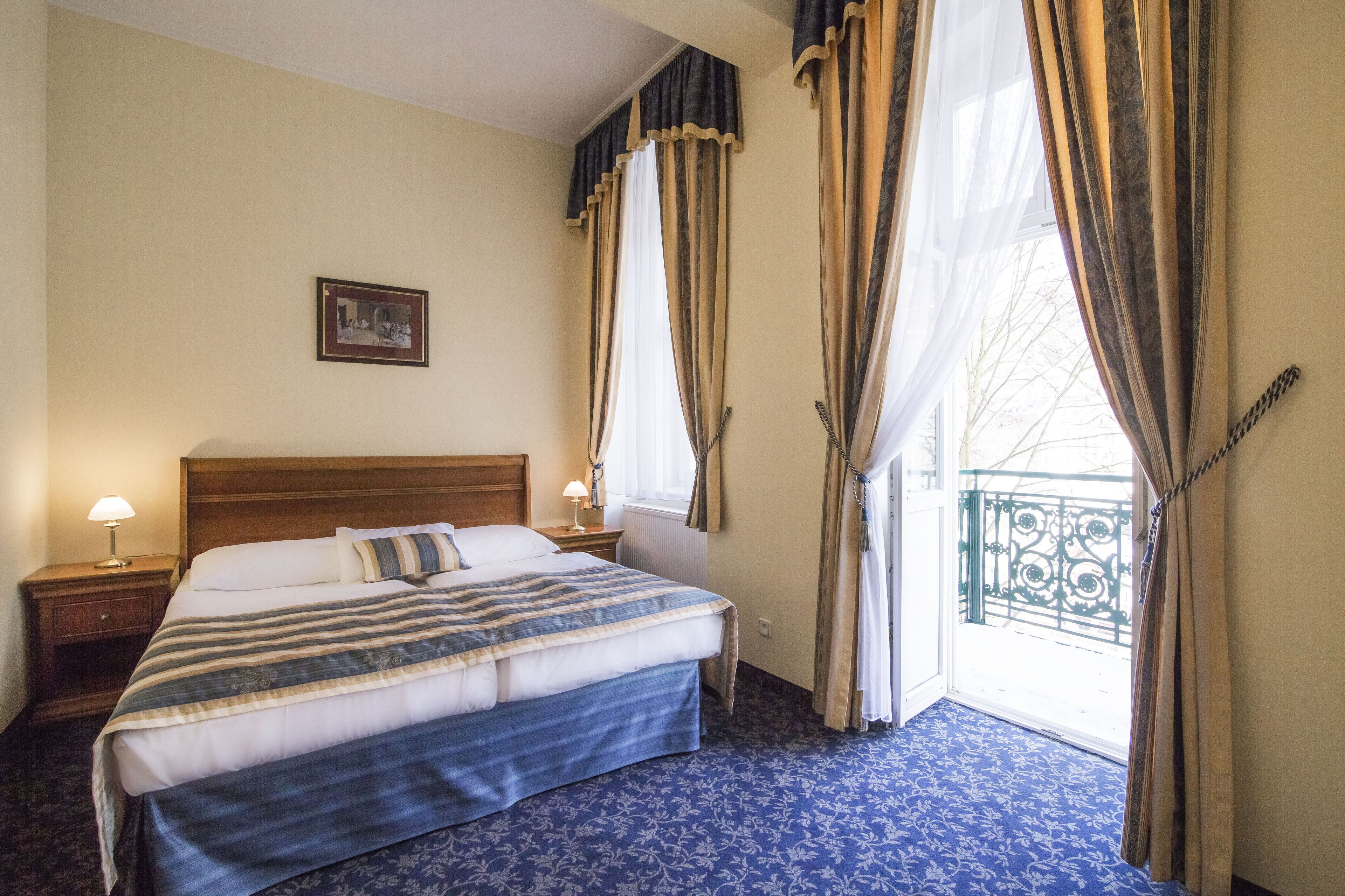 A hotel room with a large bed, a nightstand with a lamp, a window with curtains, and a view of the outside balcony.