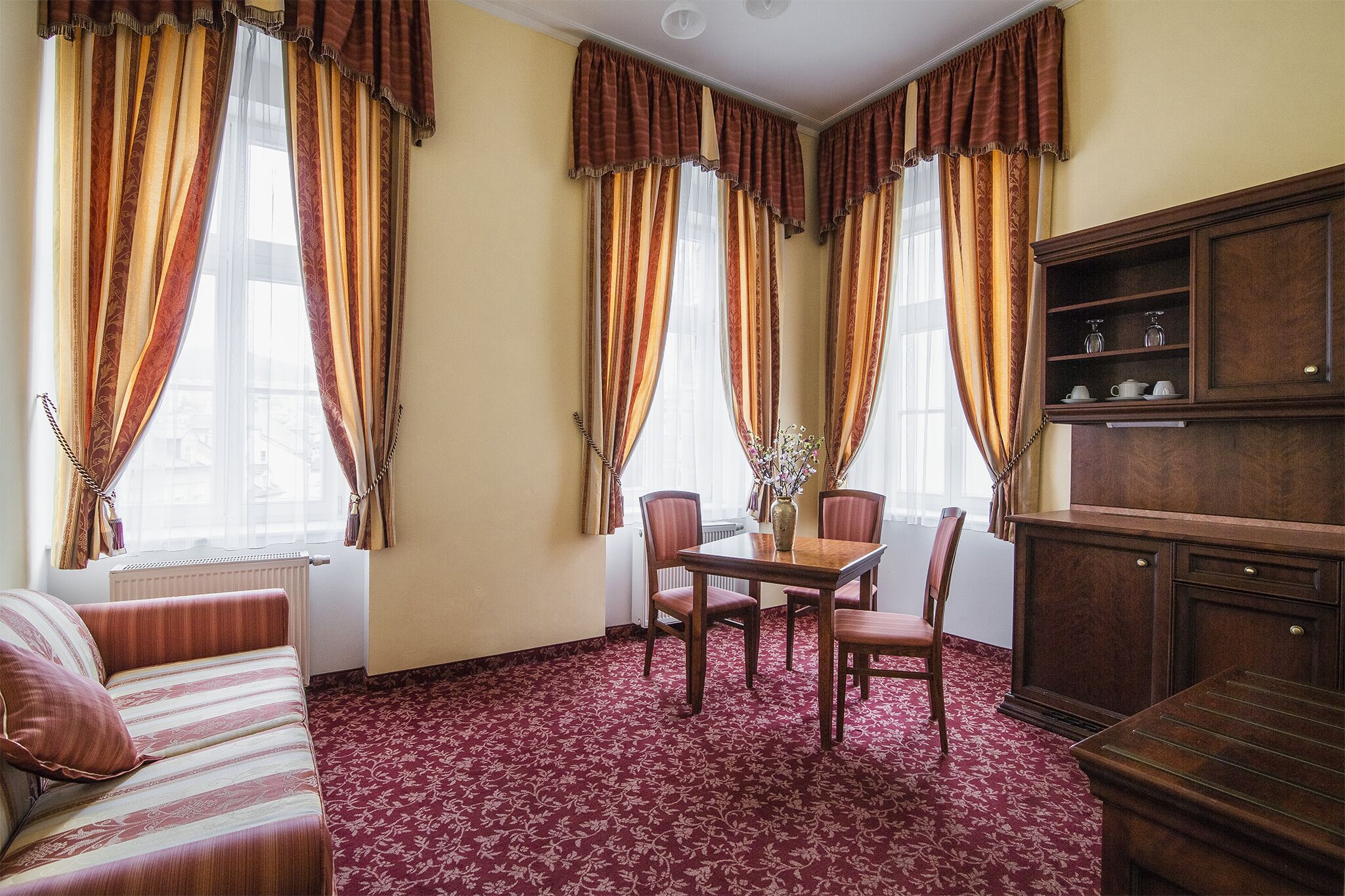 Una habitación con sofá, una mesa de comedor con sillas, un armario y ventanas grandes con cortinas.