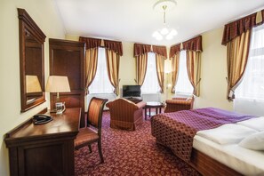 Une chambre d’hôtel avec un lit, un bureau avec une lampe, une chaise, une télévision et une fenêtre avec des rideaux.