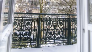 Ein Balkon mit Schnee bedecktem Boden, einem Metallgeländer und Blick auf Gebäude und Bäume im Hintergrund.