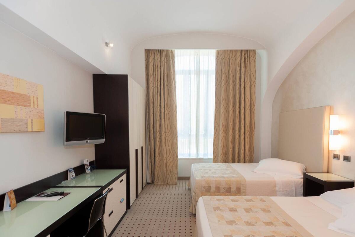 A hotel room with a desk, a flat-screen TV, two beds, a window with curtains, and a wall-mounted light.