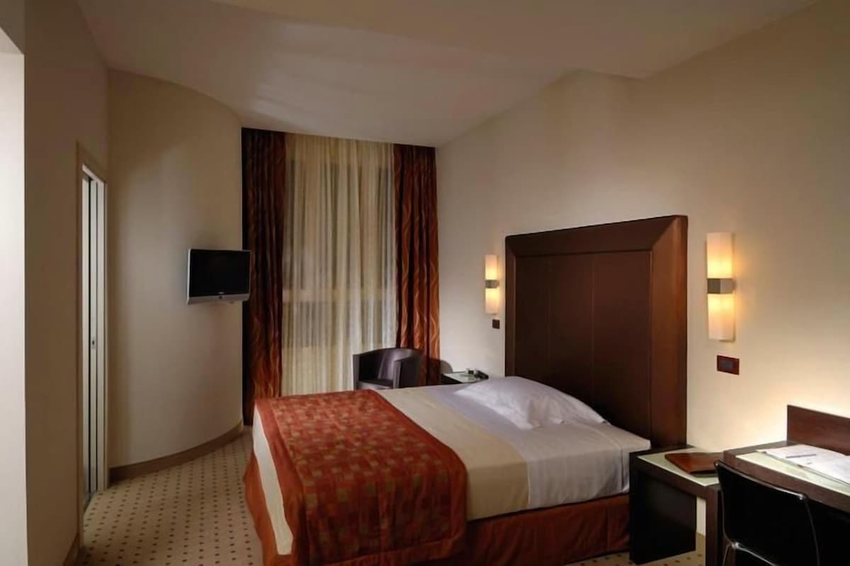 Habitación de hotel con cama, escritorio, silla, televisor y ventana con cortinas.