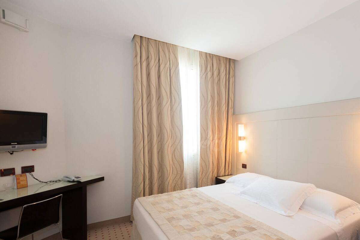 A hotel room with a bed, a desk with a television, and a large window with curtains.