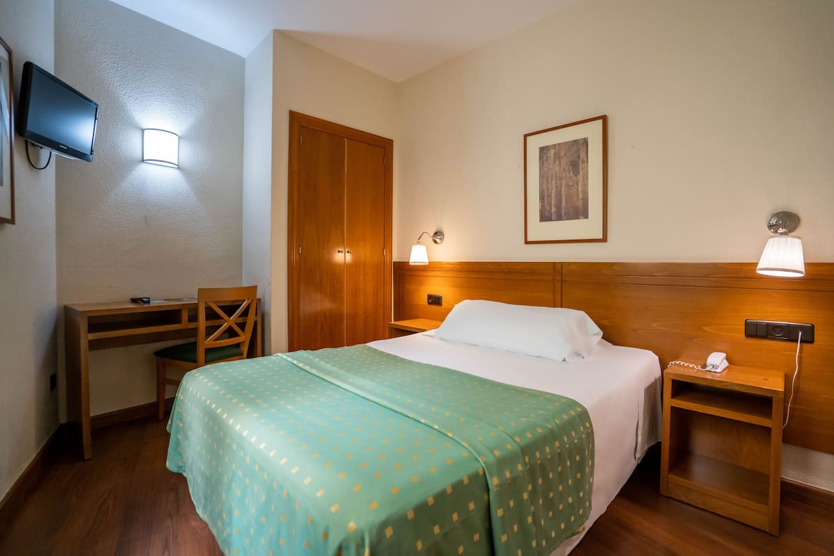 A hotel room with a bed, a desk, a chair, a TV, and a bedside table with a lamp.