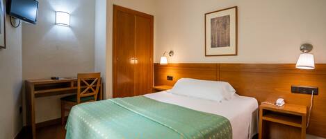 A hotel room with a bed, a desk, a chair, a TV, and a bedside table with a lamp.