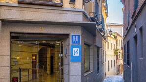 Entrada de hotel con puerta de cristal, un letrero azul que indica los servicios y una calle empedrada.