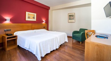 Quarto de hotel com cama de madeira, uma poltrona verde, uma escrivaninha com televisão de tela plana e duas imagens emolduradas nas paredes.